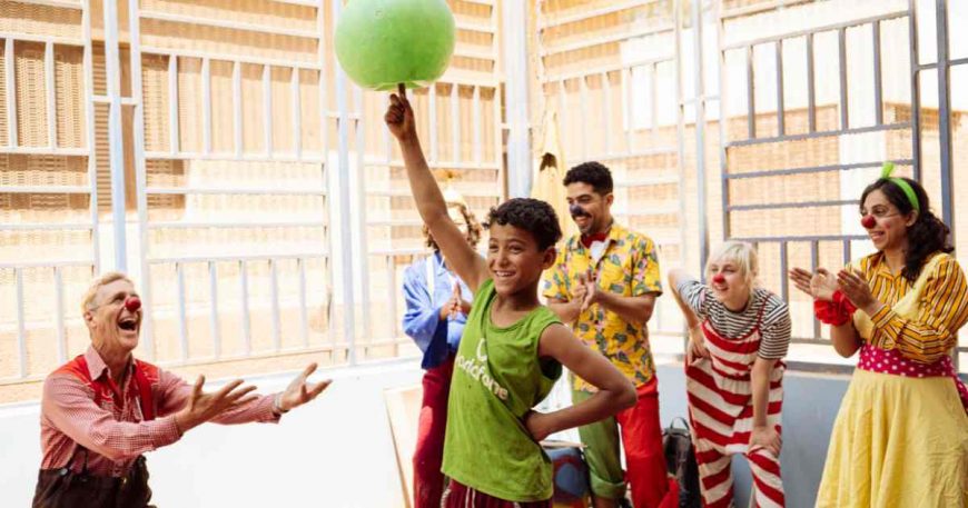 Kid balancing a ball in front of a group of clowns