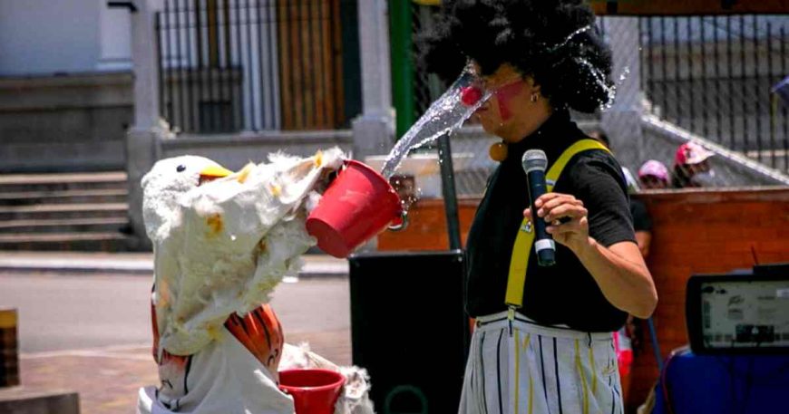 A clown dressed as a chickenn throws a bucket of water in the face of another clown.