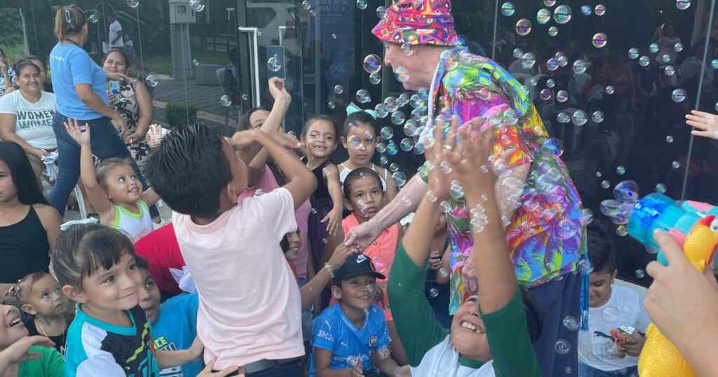 A clown in a brightly colored shirt and hat stands among children and bubbles.