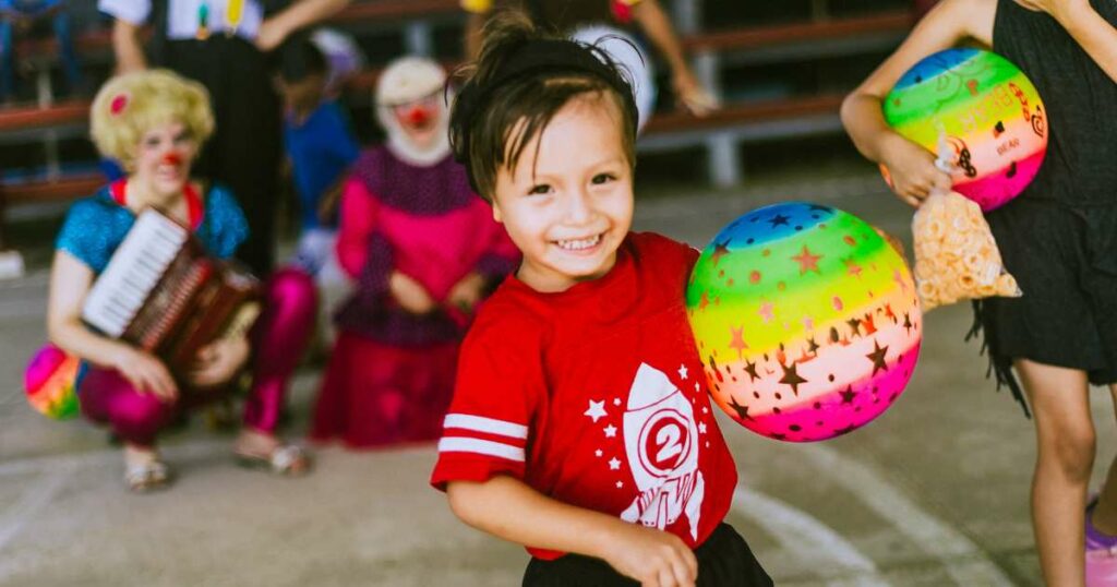 A boy in a red shirt holds a rainbow colored ball at a clown show.