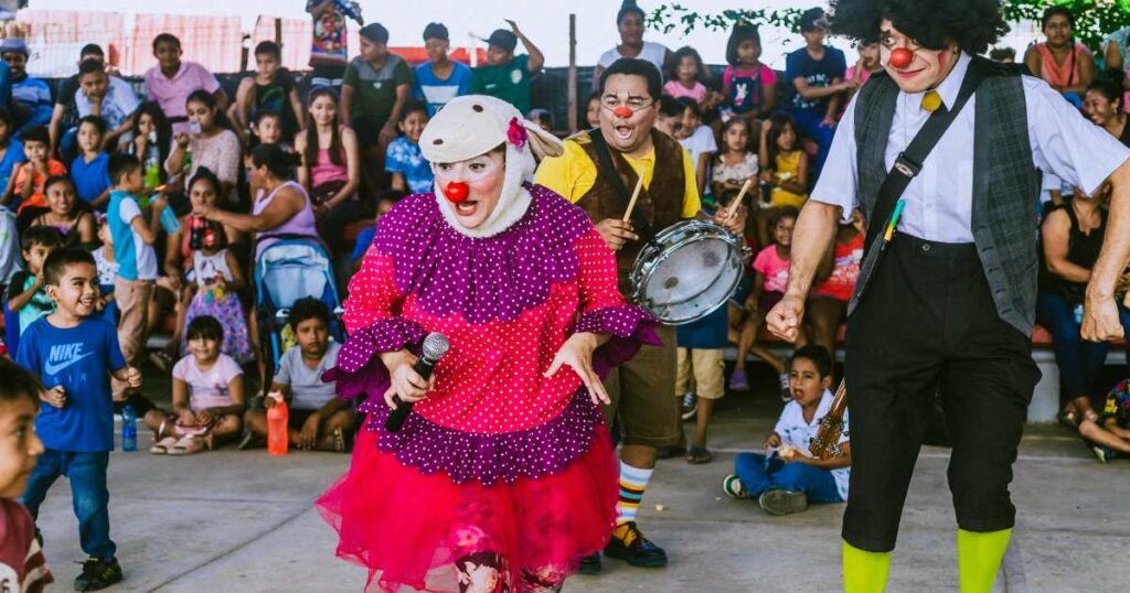 A clown in a bright pink dress and a lamb hat invites children to the stage.